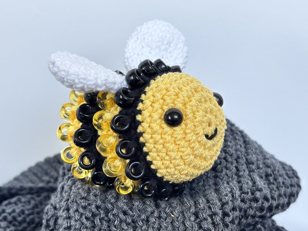 Close-up of a crocheted bee amigurumi with yellow crochet, black and yellow bead loops, white wings and black bead eyes on grey knit fabric.
