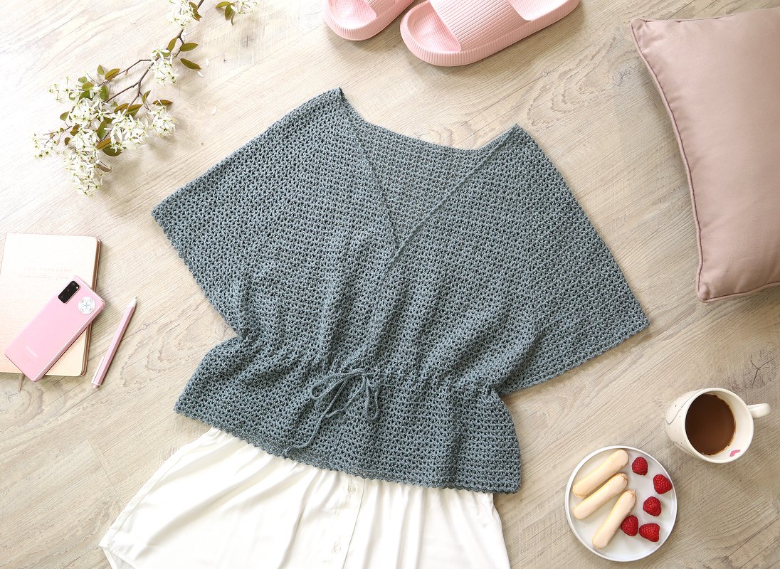 Overhead view of a dusty blue crocheted kimono-style top with a V-neck and drawstring waist laid on a light wood floor with lifestyle accessories.