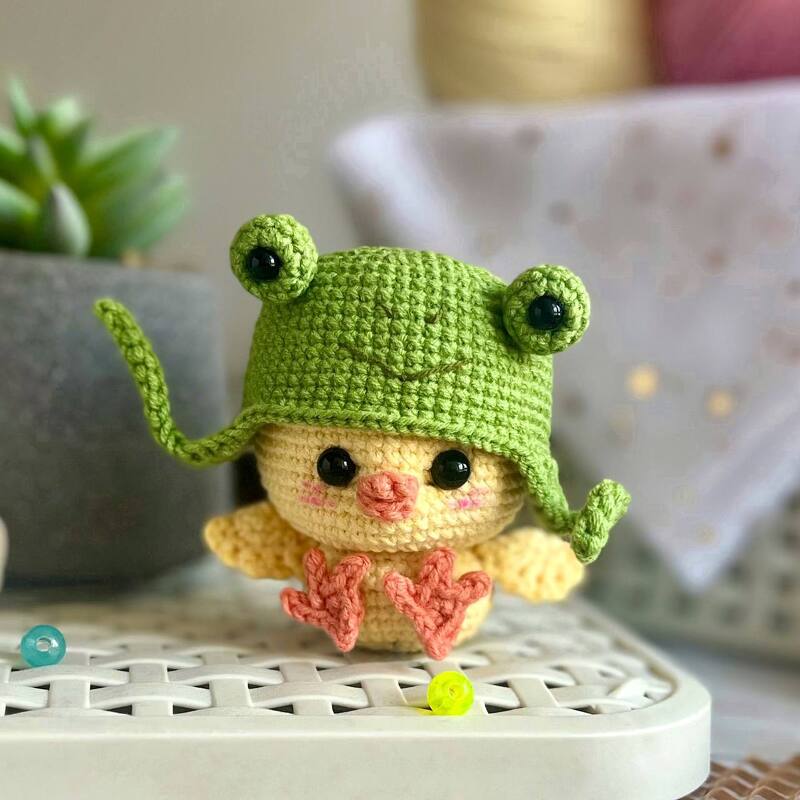 Close-up of a yellow crocheted chick toy wearing a green frog-shaped hat, sitting on a white surface.