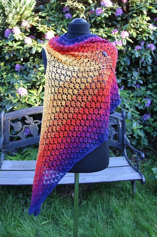 Mannequin wearing an airy diagonal gradient lace poncho-shawl in purple, red and yellow tones, photographed outdoors against a garden backdrop.