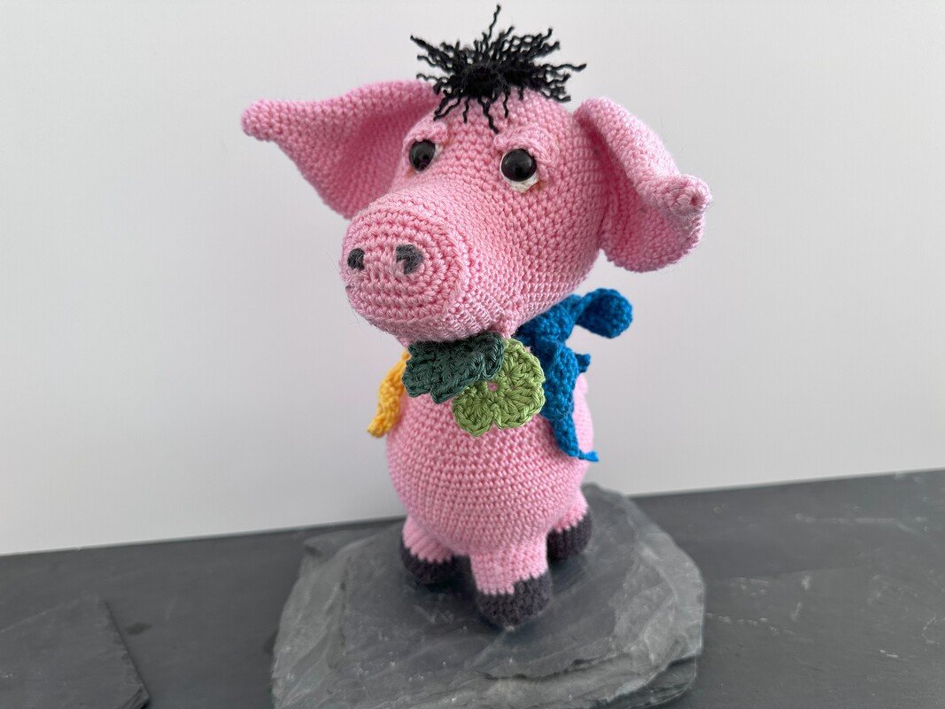 Rosa gehäkeltes Amigurumi-Schwein mit schwarzen Locken und buntem Blätterkranz, auf einem dunklen Stein.