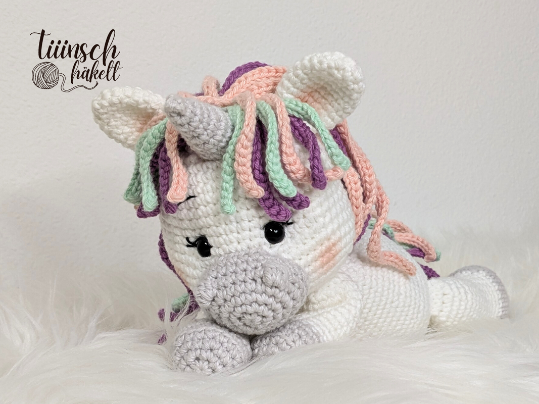 Weißes gehäkeltes Einhorn-Amigurumi mit pastellfarbener Mähne, Horn und schwarzen Augen, liegend auf flauschigem Untergrund