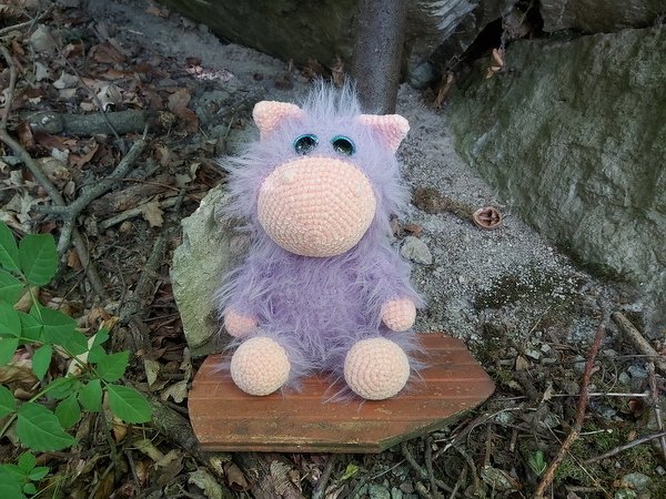 Gehäkeltes flauschiges Amigurumi-Tier in Lila mit pfirsichfarbener Schnauze, sitzend auf Holz