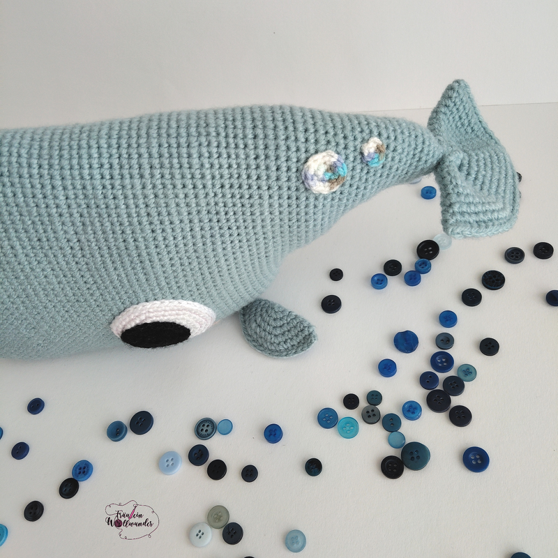 Hellblau gehäkelter Wal Amigurumi liegend, umgeben von blauen Knöpfen