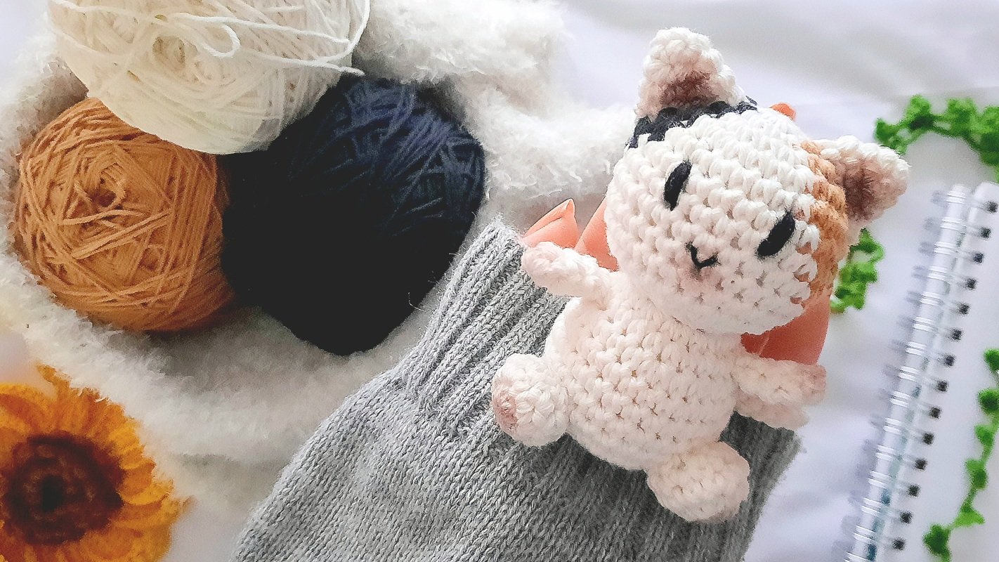 Close-up of a white, orange, and black crocheted amigurumi cat doll next to three balls of yarn, a grey knitted item, and a yellow crochet flower.