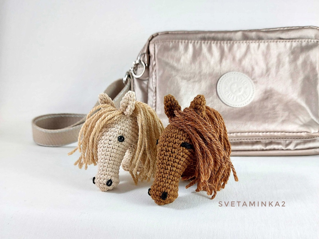 Two crocheted horse head keychains, one beige and one brown, with yarn manes, placed in front of a light-colored bag.
