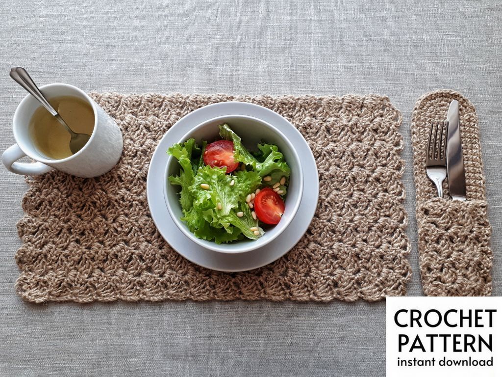 Top-down view of a crocheted placemat and matching cutlery holder in natural beige yarn with a salad bowl and mug on a light grey tablecloth.