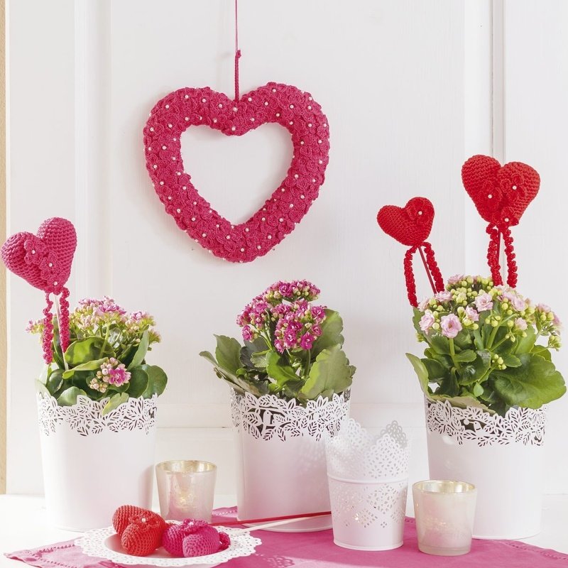 Gehäkelte Herzdeko in Pink und Rot mit blühenden Pflanzen in weißen Spitzenübertöpfen
