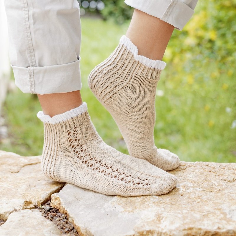 Beigefarbene, gestrickte Damensocken mit Lochmuster und weißem Rüschenrand an den Füßen einer Person auf einem Steinuntergrund.