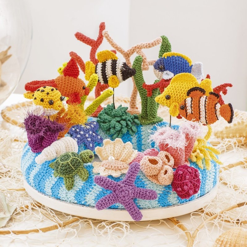 Gehäkeltes Korallenriff mit Fischen, Schildkröte, Seesternen und Muscheln auf blau-weiß gestreiftem Sockel.