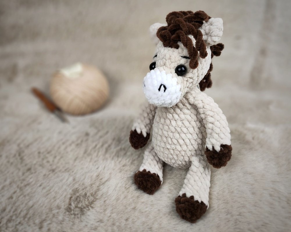 Nahaufnahme eines beigen, gehäkelten Amigurumi-Pferdes mit brauner Mähne und dunklen Hufen auf hellem, flauschigem Untergrund