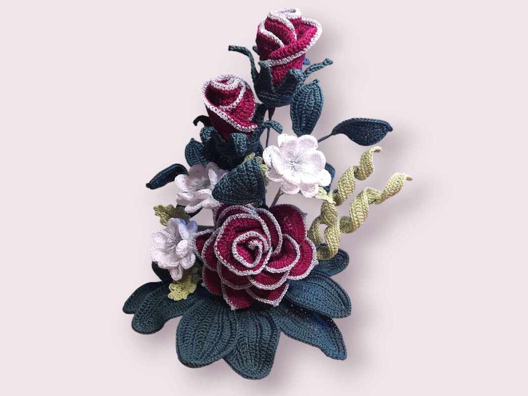 Gehäkeltes Blumenarrangement mit roten Rosen, weißen Blüten und grünen Blättern auf hellrosa Hintergrund