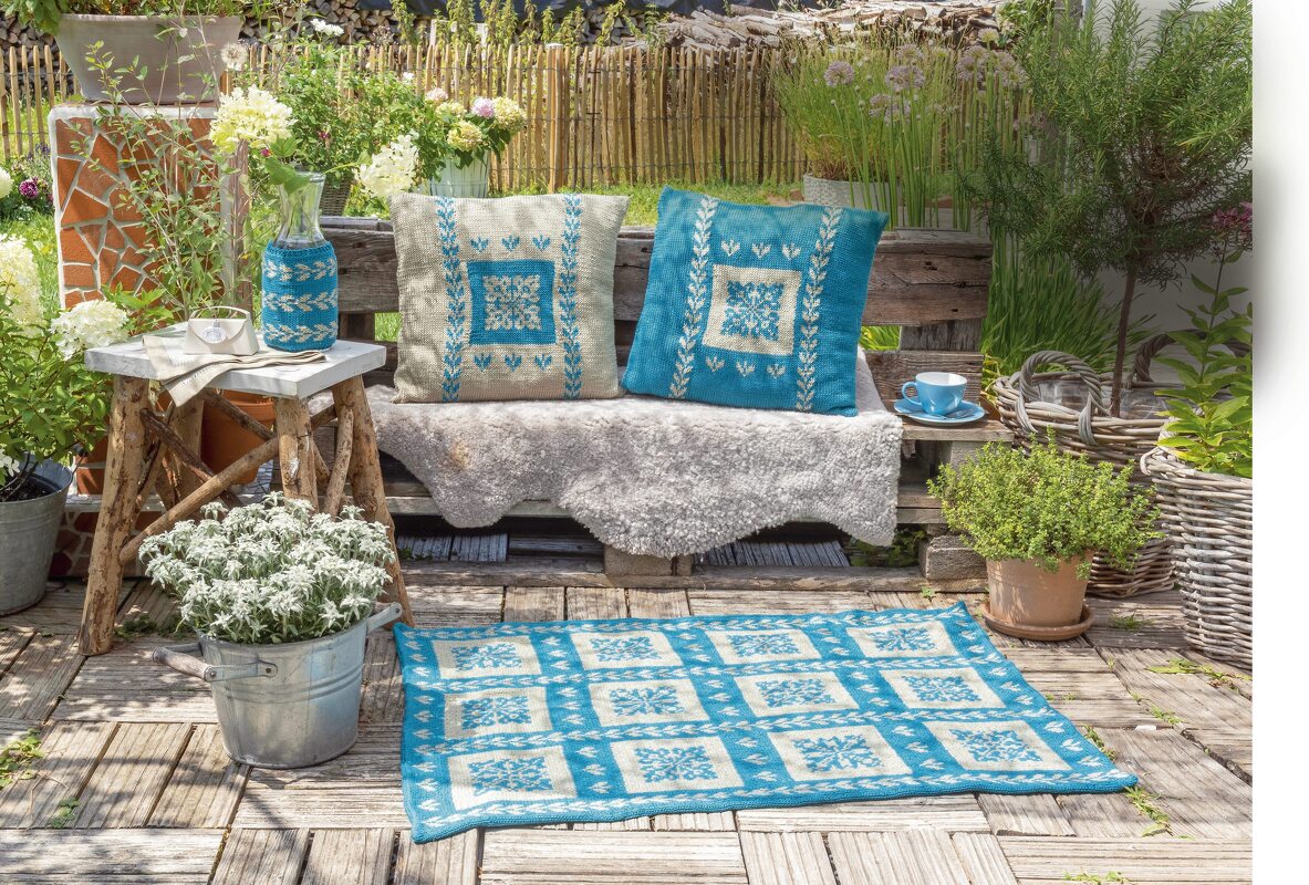 Zwei gestrickte Kissen, eine gestrickte Vasenhülle und ein gestrickter Teppich in Blau und Creme mit traditionellen Mustern auf einer Gartenbank