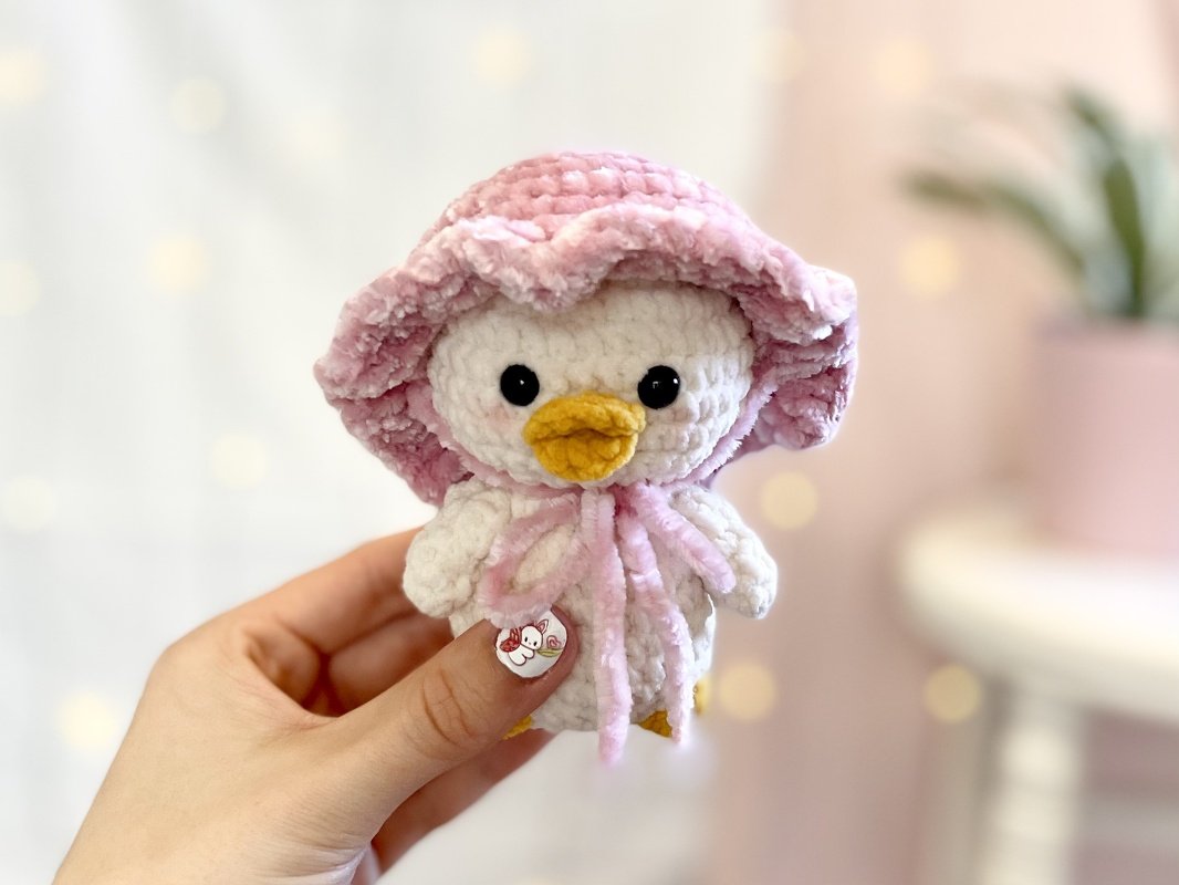 Hand hält ein weißes gehäkeltes Amigurumi-Küken mit rosa Häubchen, gelbem Schnabel und dunklen Augen.