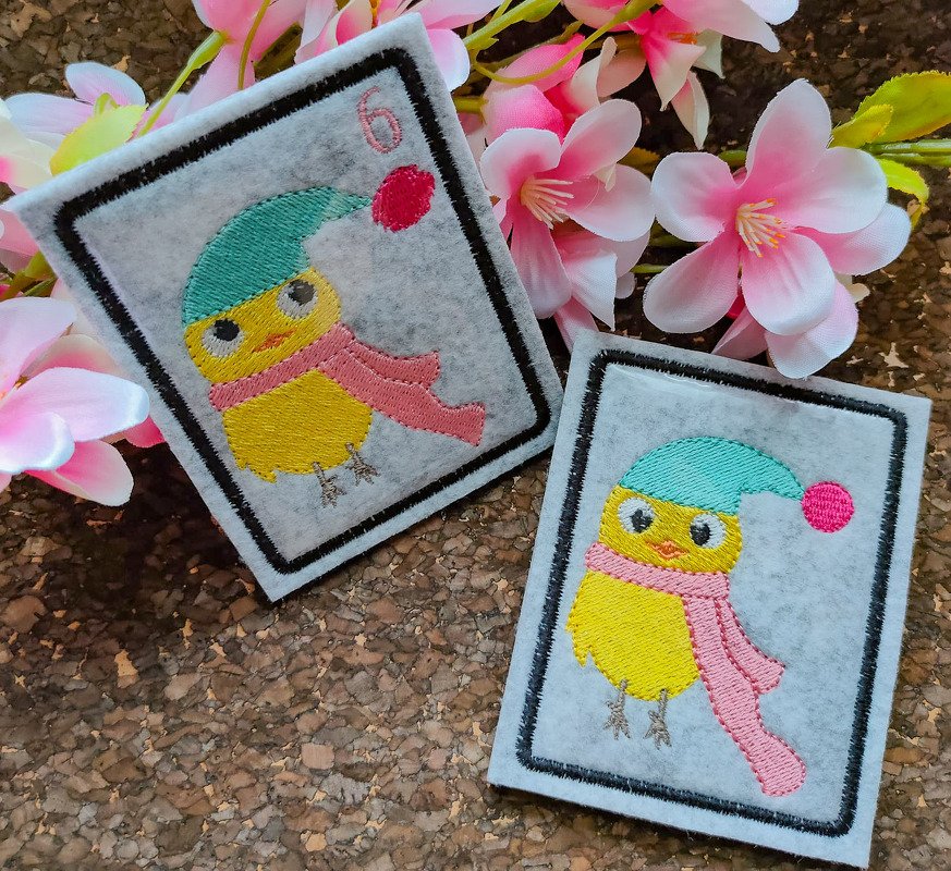Zwei hellgraue Filzquadrate mit gesticktem gelben Küken, türkisfarbener Mütze, rosa Schal und schwarzer Randstickerei; auf einer Karte die Zahl 6
