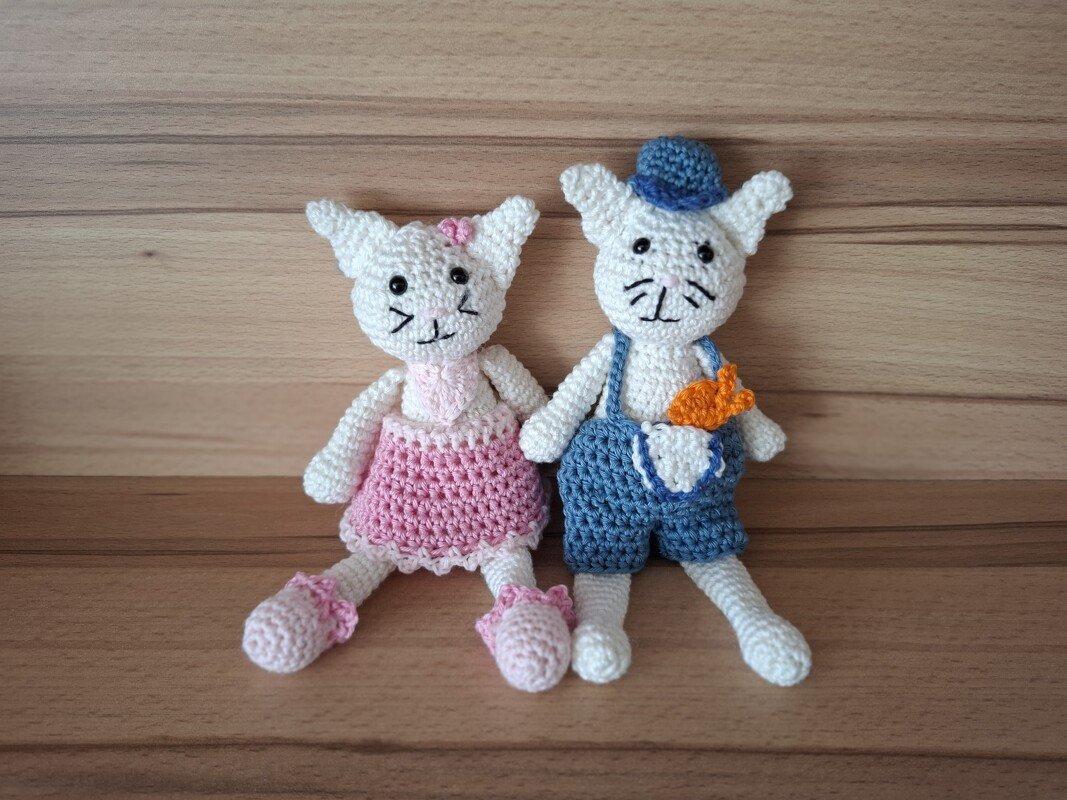 Zwei gehäkelte Katzen-Amigurumi nebeneinander, links rosa Kleid, rechts blaue Latzhose mit Hut und orangefarbener Karotte in der Tasche
