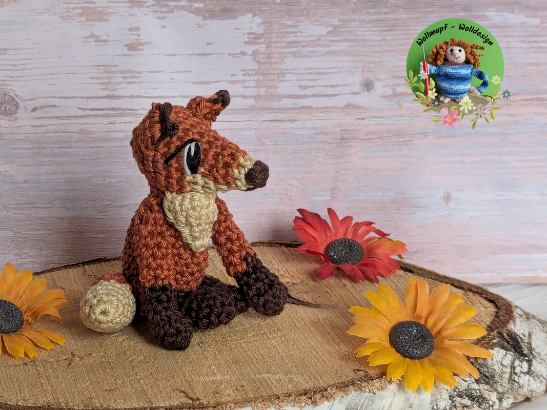 crocheted amigurumi fox in rust, cream and dark brown yarn seated on a wooden slice with artificial sunflowers