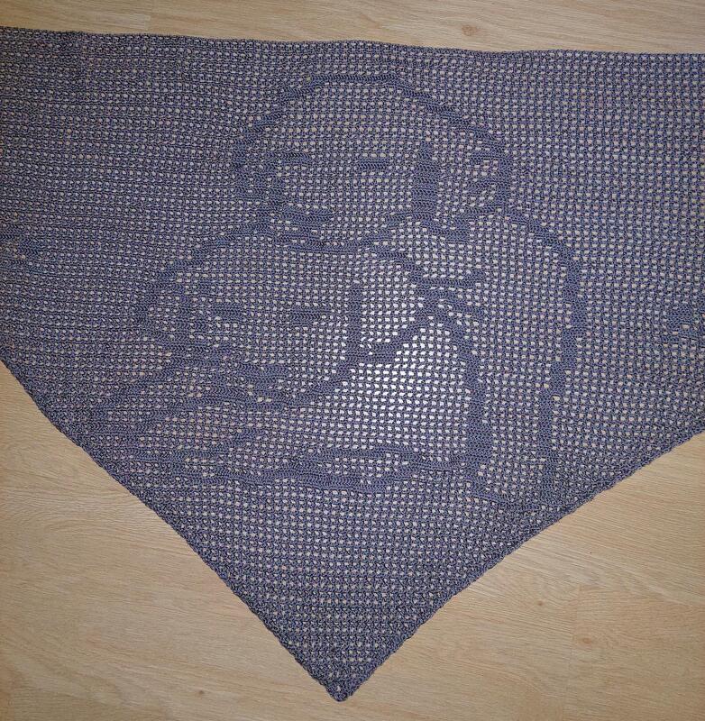 Gehäkeltes dreieckiges Tuch in Dunkelgrau mit eingearbeitetem figürlichem Motiv, liegend auf Holzboden.