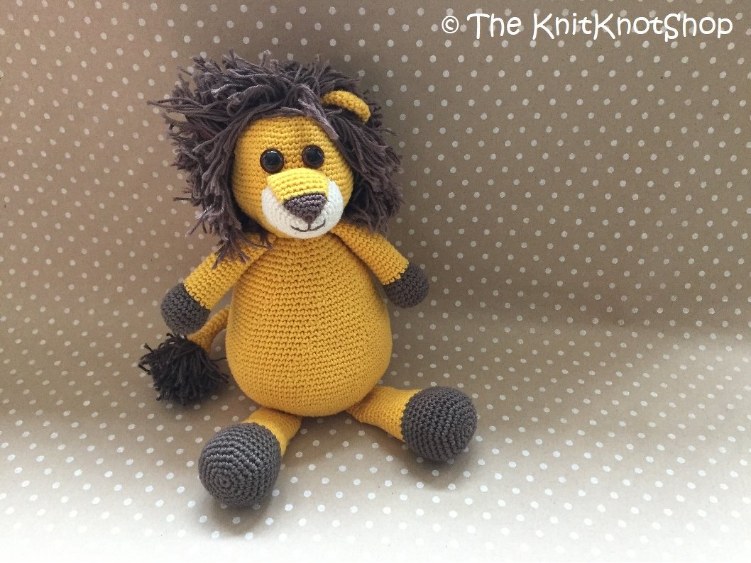 Gehäkelter Amigurumi-Löwe in Senfgelb mit dunkelbrauner Mähne und Schwanzquaste, sitzend vor gepunktetem Stoff