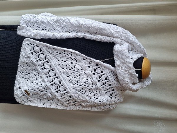 Weißer gehäkelter Schal mit diagonalen Reihen und blütenartigen Lochmustern, drapiert; kleine holzähnliche Applikation sichtbar.