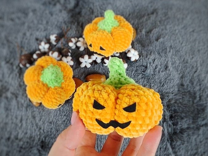 Drei gehäkelte orangefarbene Kürbisse, einer davon mit schwarzem Halloween-Gesicht, einer wird in der Hand gehalten.