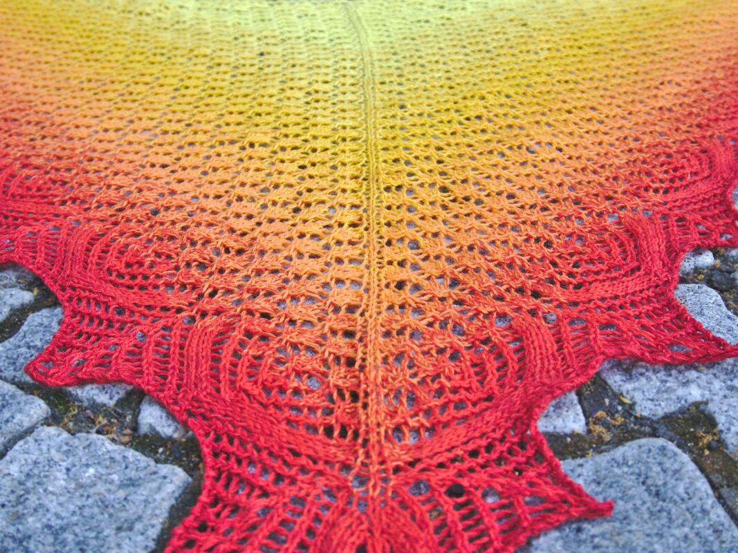 Triangular yellow-to-red gradient lace shawl with scalloped leaf border and center spine laid flat on stone.