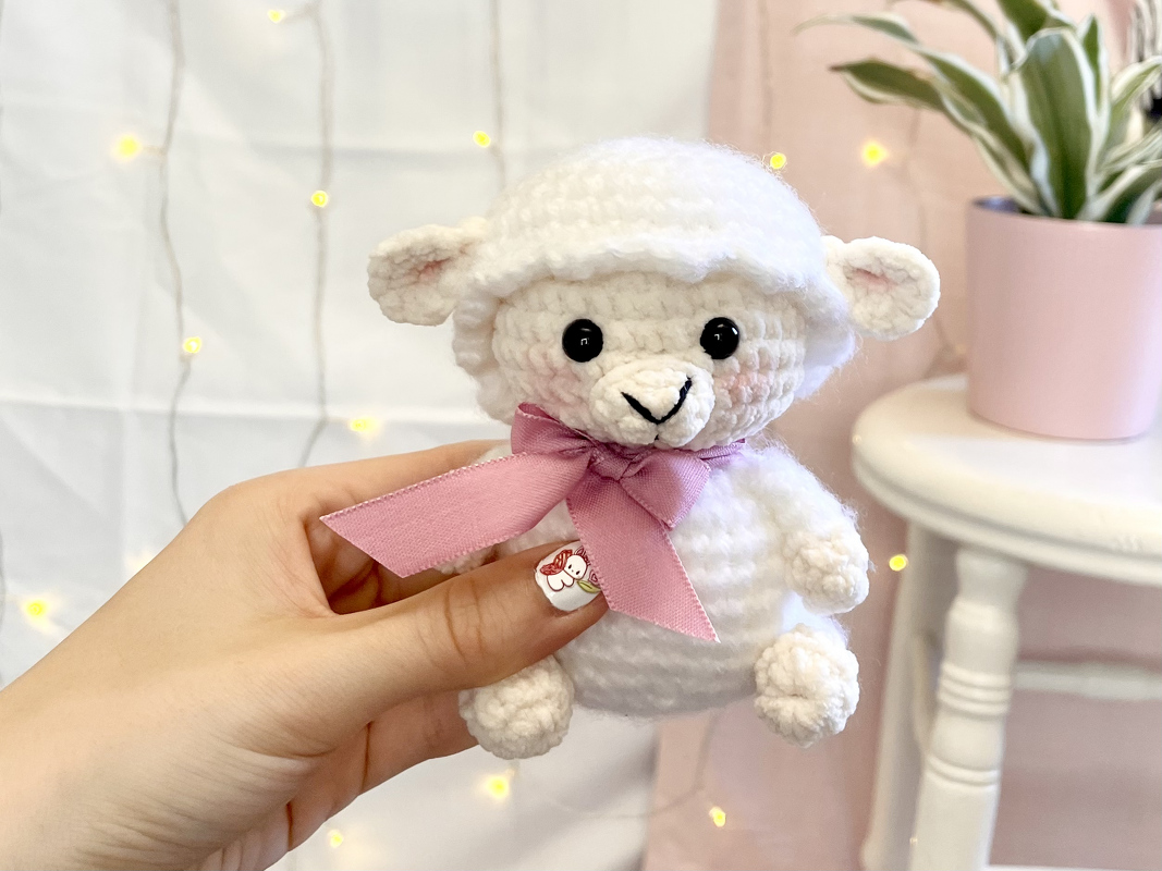 Weißes gehäkeltes Amigurumi-Schaf mit rosa Schleife, in der Hand gehalten