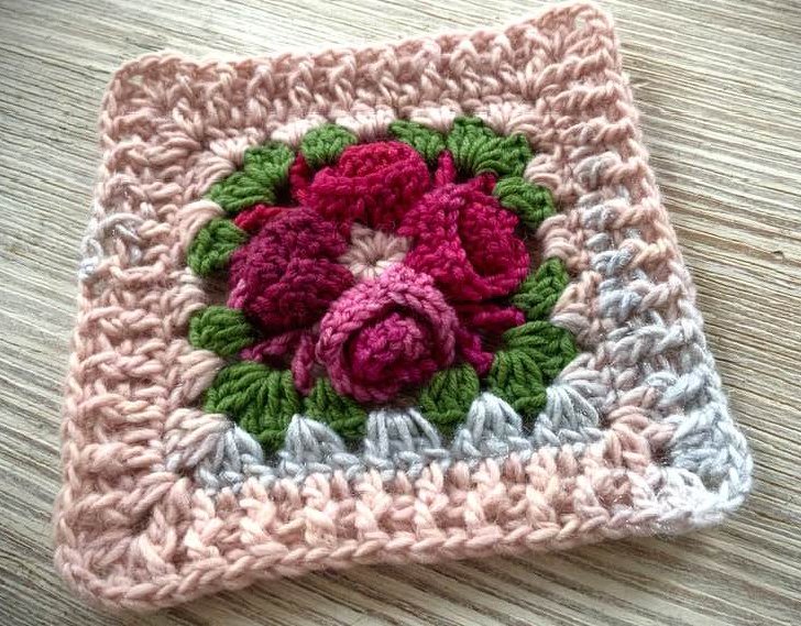 Handgehäkeltes Granny Square mit plastischen roten Rosen, grünen Blättern und zartrosa-grauem Rand