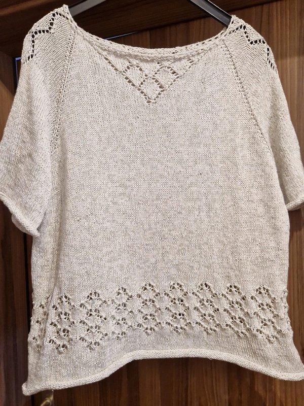 Helles gestricktes Damen-Kurzarmshirt mit Lochmuster am Saum und Rautendetail am Ausschnitt, auf einem Bügel hängend.