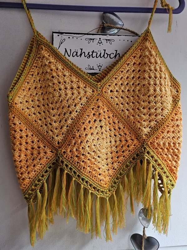 Handgehäkeltes Damen-Top in Orange und Gelb mit grünen Akzenten und Fransen, an einem Kleiderbügel hängend.