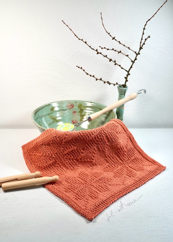 Terrakottafarbenes gestricktes Spültuch mit erhabenem Blattmuster auf hellem Tisch, im Hintergrund Keramikschale und Vase mit Zweig