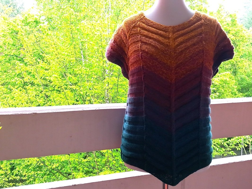 Gestricktes Damen-Kurzarmshirt mit V‑Chevron‑Muster und Verlaufsfarben von Senfgelb bis Petrol auf Schneiderpuppe