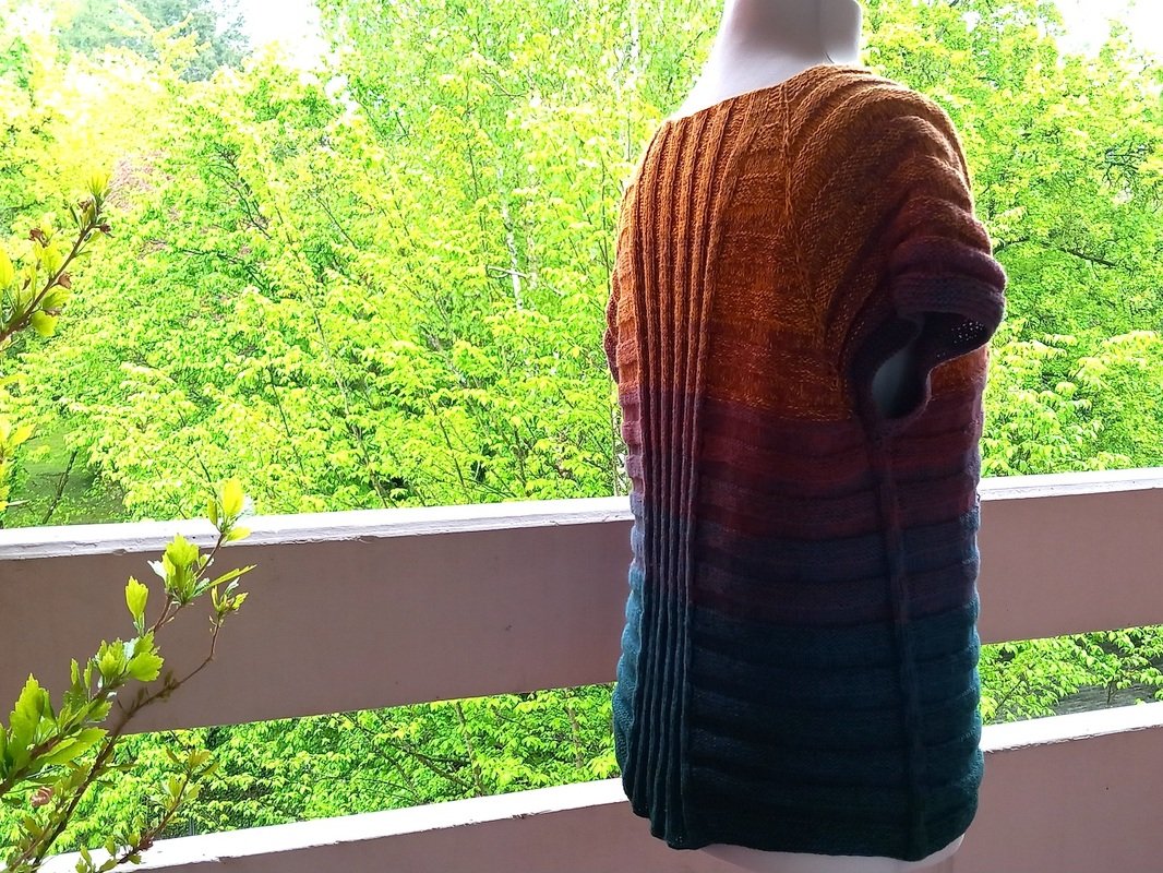 Mannequin mit gestrickter Tunika, Farbverlauf von Braun-Orange zu Blau, Rückenansicht auf Balkon vor grünen Bäumen