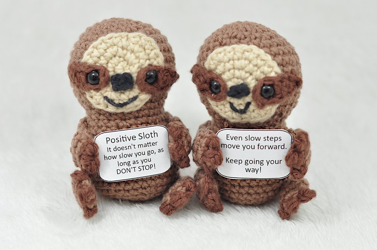 Zwei gehäkelte Amigurumi-Faultiere in Braun- und Beigetönen halten kleine Schilder mit aufmunternden Sprüchen.