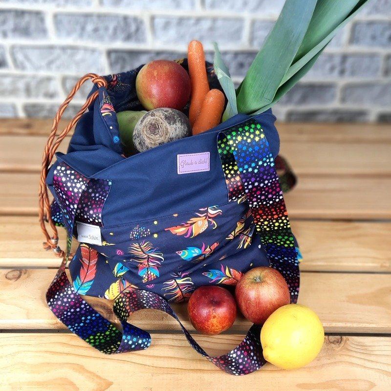 Dunkelblaue Stofftasche mit buntem Federmuster und regenbogenfarbenen Trägern, gefüllt mit Lauch, Karotten, Äpfeln, Roter Bete und einer Zitrone auf einer Holzoberfläche.