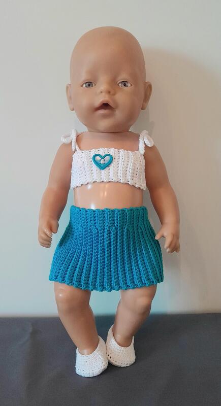 Babypuppe mit weißem gehäkeltem Top mit blauem Herz, blauem Faltenrock und weißen Schühchen