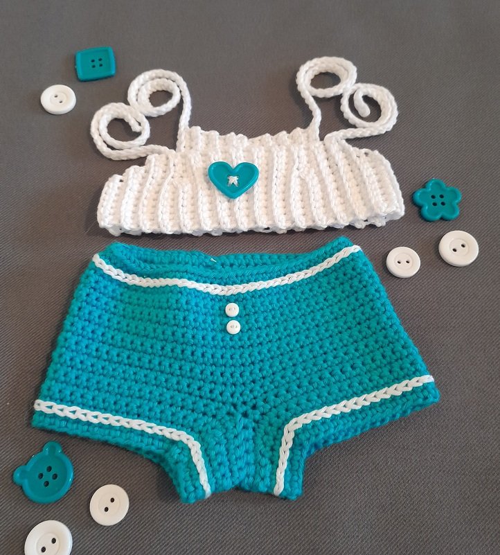 Gehäkeltes Puppenkleidungs-Set in Türkis und Weiß auf grauem Hintergrund mit Oberteil, Hose und dekorativen Knöpfen.