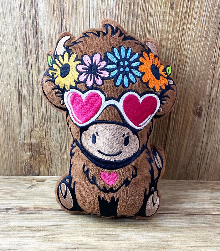 gestickte braune Rinder-Figur mit buntem Blumenkranz und pinken herzförmigen Sonnenbrillen, Vorderansicht auf Holzuntergrund