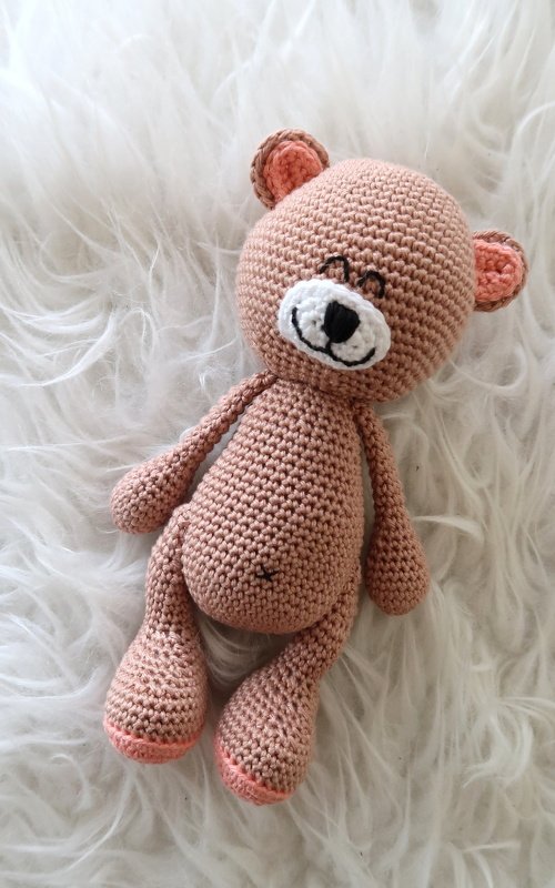Gehäkelter hellbrauner Amigurumi-Teddybär mit weißer Schnauze und rosa Ohren liegt auf weißem Fell