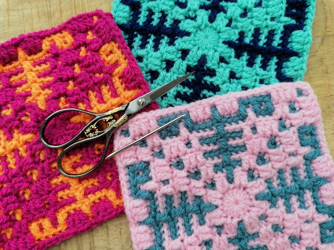 Drei mehrfarbige gehäkelte Granny Squares mit Schere und Metallnadel auf einer Holzoberfläche