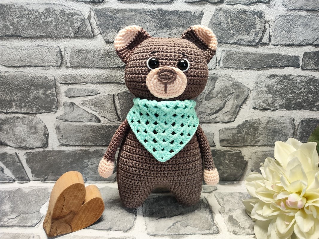 Brauner Amigurumi-Teddybär mit mintgrünem Halstuch und beigen Details vor grauer Ziegelwand
