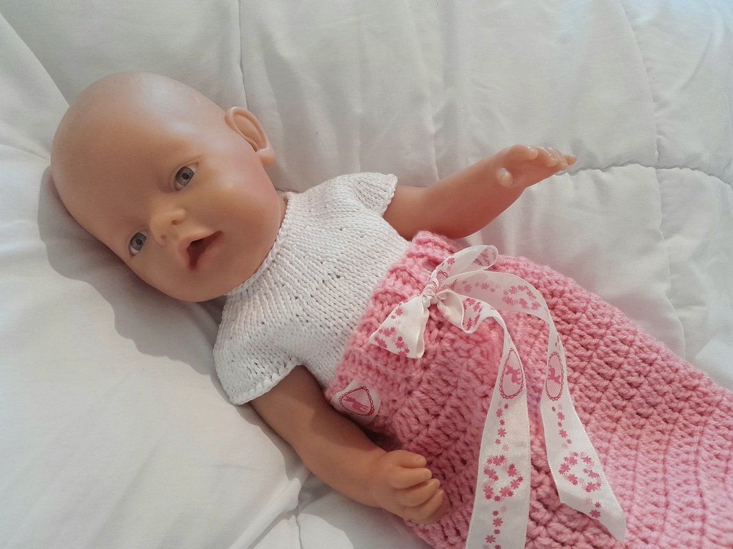 Babypuppe liegt auf weißer Decke, trägt weißes gestricktes Oberteil und rosa gehäkelten Rock mit Band