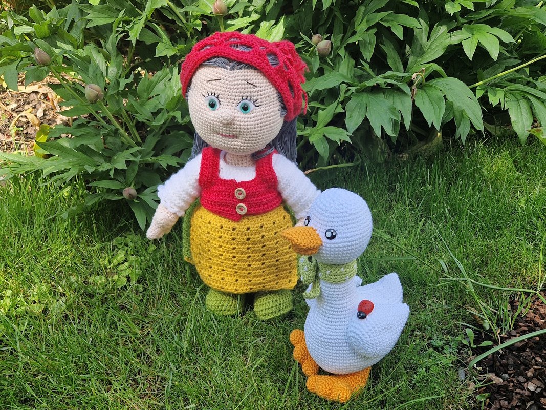 Gehäkelte Amigurumi-Puppe mit rotem Kopftuch neben weißer Häkelgans auf grünem Rasen.