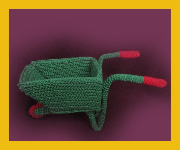 Green crocheted wheelbarrow with red handles and wheel on a purple background.