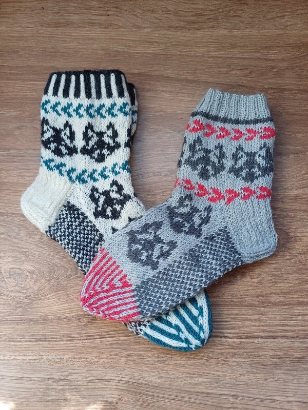 Zwei gestrickte Socken mit Tiermotiven in Grau, Weiß, Rot und Türkis auf Holzhintergrund.
