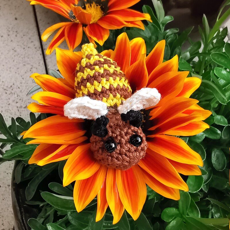 Gehäkelte Amigurumi-Biene mit gestreiftem Hinterleib und weißen Flügeln auf orange-gelber Blüte