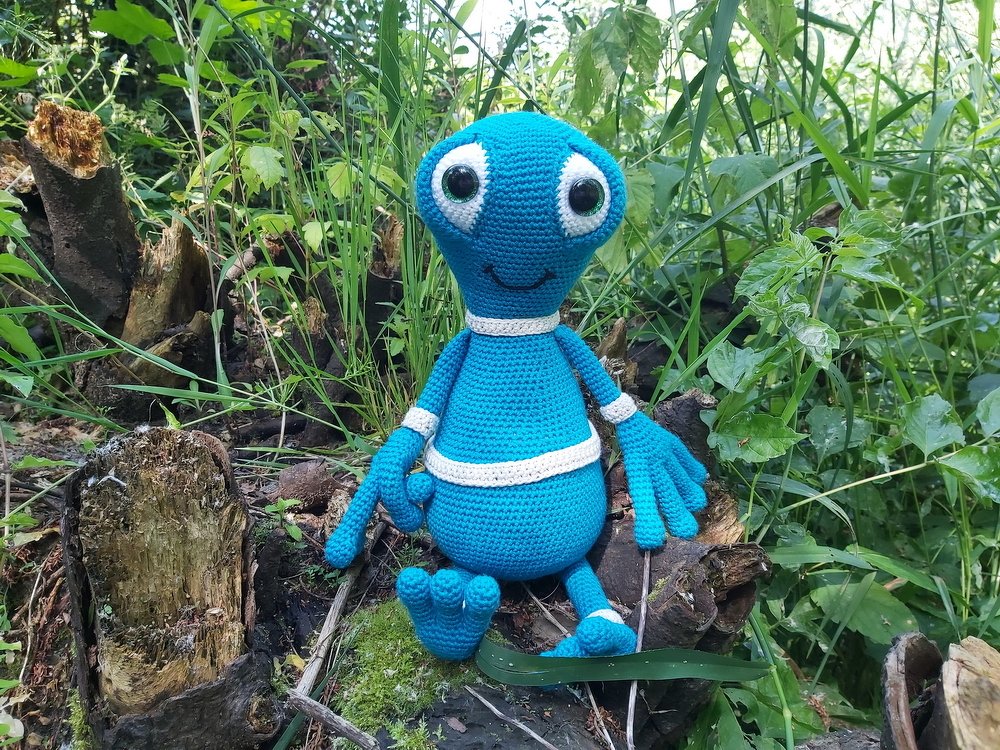 Blaues gehäkeltes Alien-Amigurumi mit großen Augen sitzt auf einem Baumstumpf im Grünen.