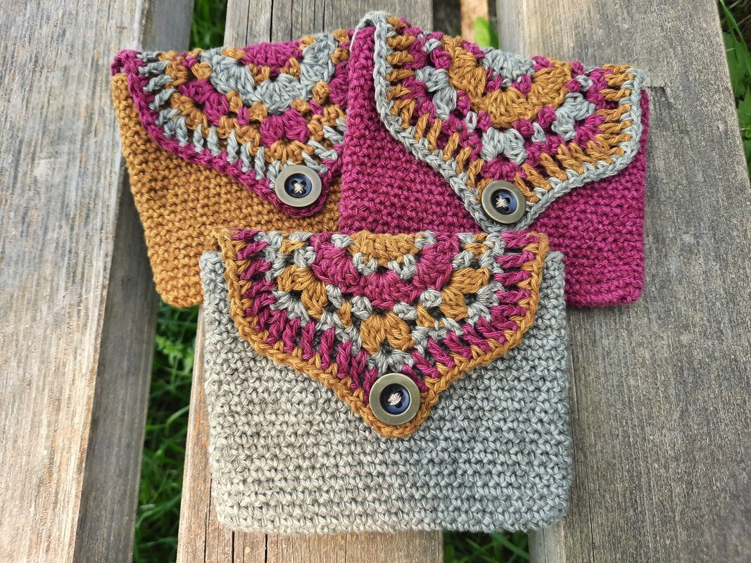 Drei gehäkelte Täschchen in Senf, Pink und Hellgrau mit mehrfarbigen Granny‑Klappen und Knopfverschlüssen auf einer Holzbank