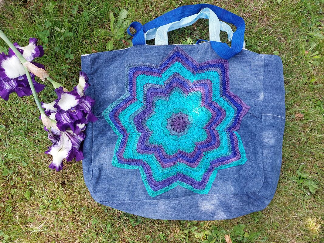 Blaue Denim-Einkaufstasche mit gehäkeltem Stern in Türkis und Lila auf Gras neben Schwertlilien