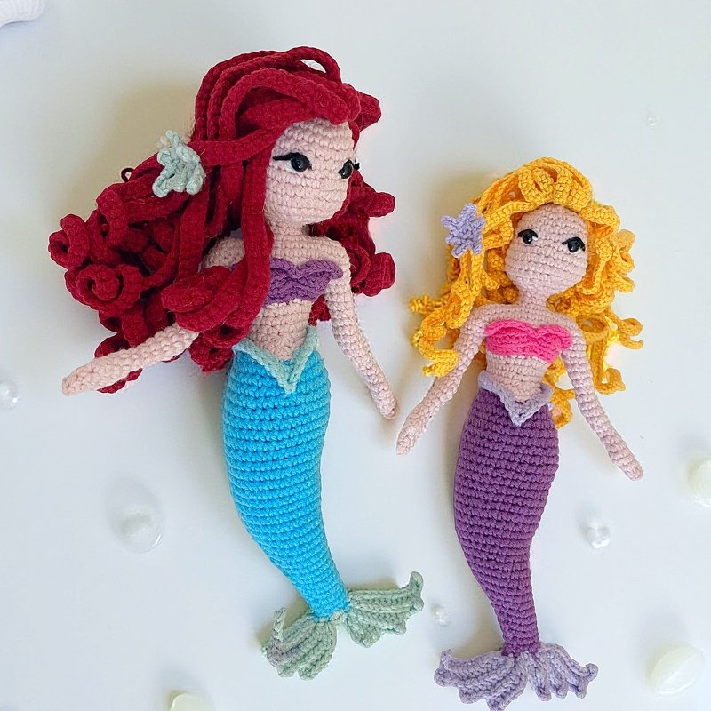 Zwei Amigurumi-Meerjungfrauenpuppen nebeneinander, eine mit roten Locken und türkisem Schwanz, die andere mit gelben Locken und violettem Schwanz